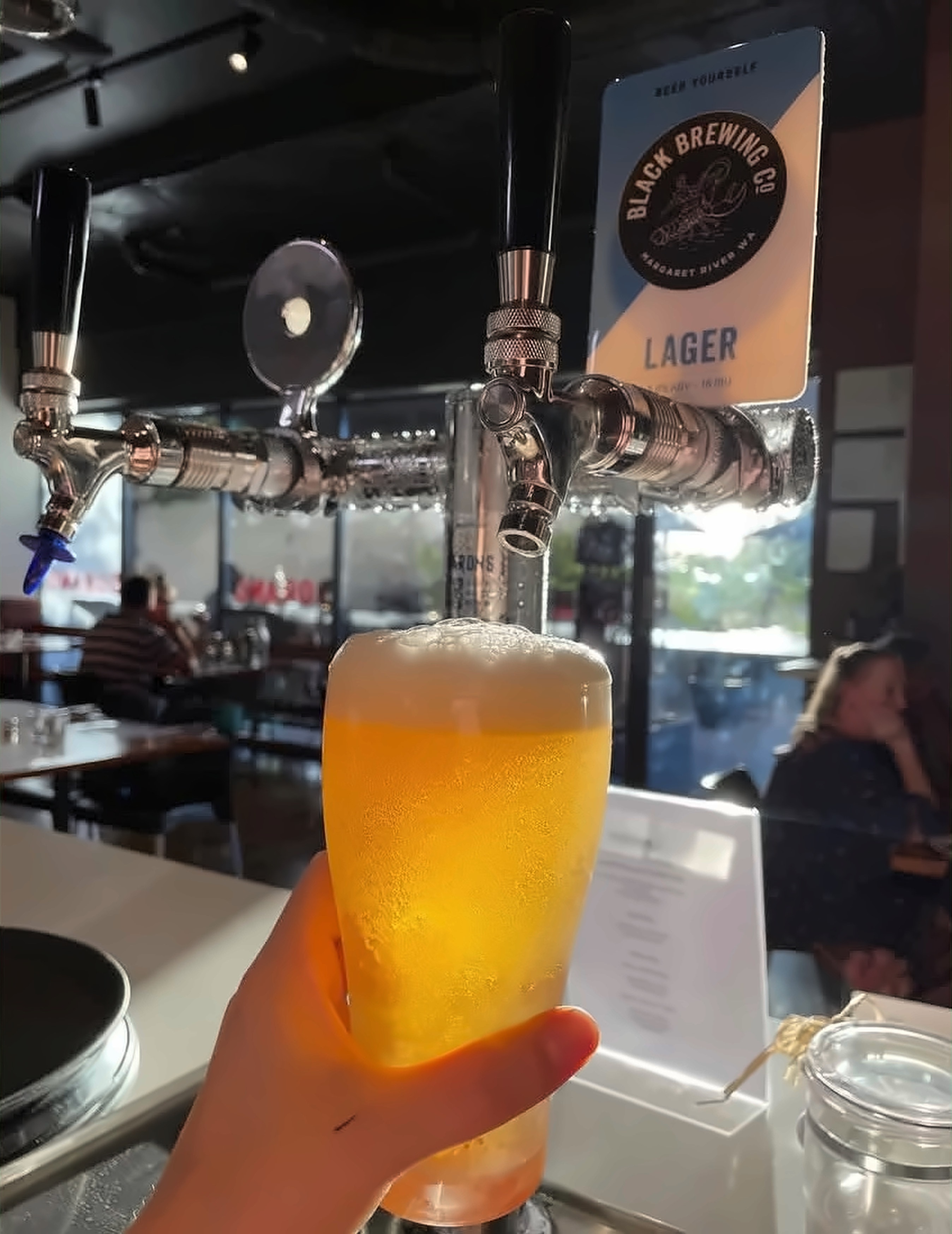 A freshly poured lager held up in front of the beer taps, with diners in the restaurant behind
