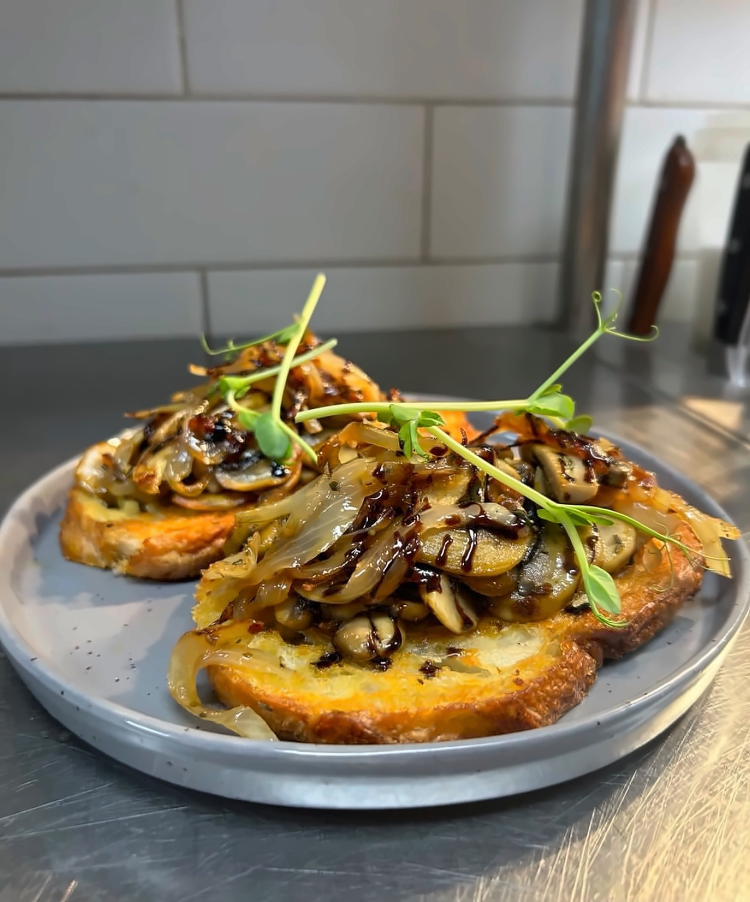 Toasted bread piled with caramelised onion and mushroom, balsamic glaze and pea shoots, on the kitchen pass