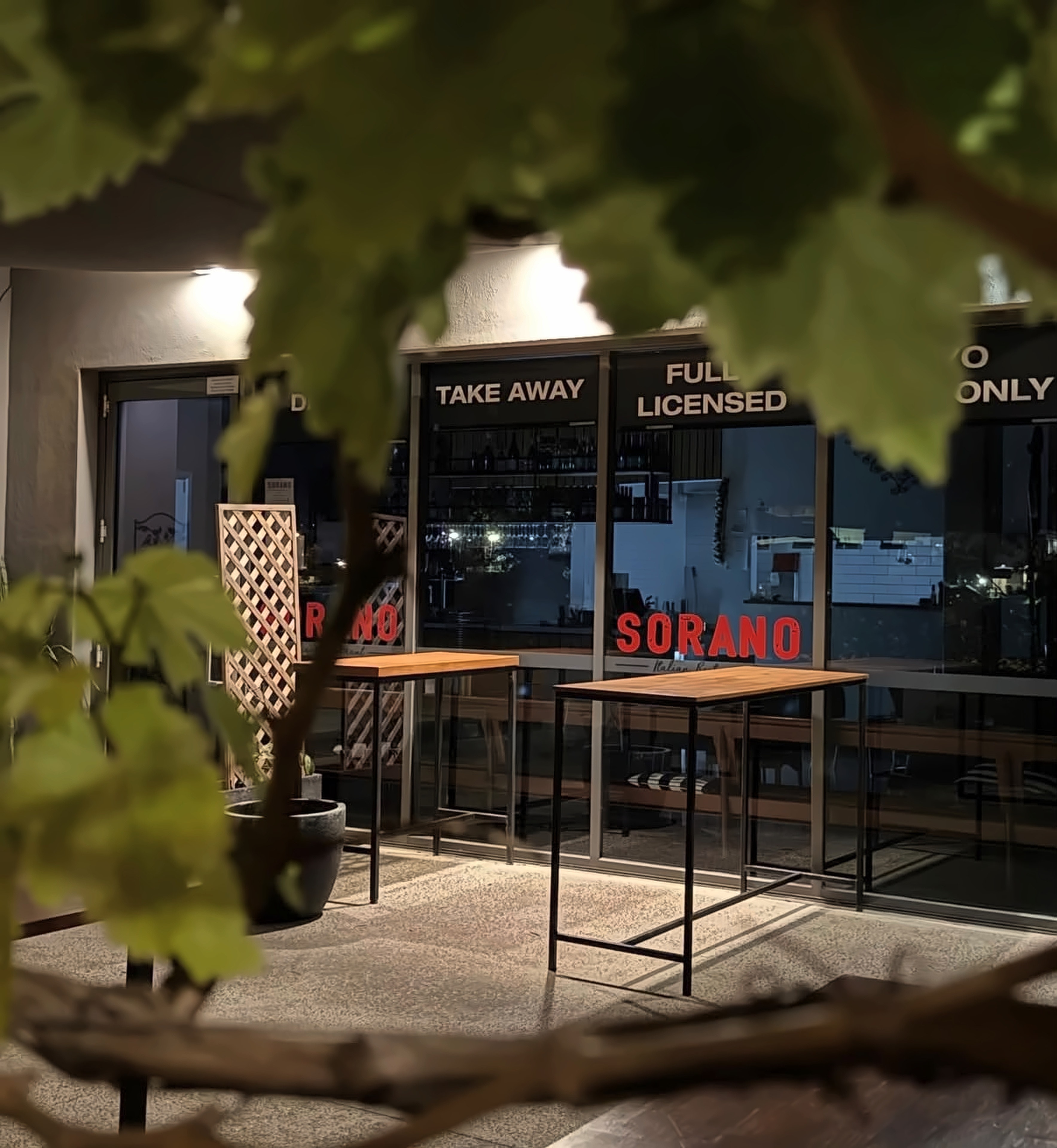 Sorano at night seen through grapevine leaves: lit windows with red SORANO lettering, timber bar tables and a lattice screen